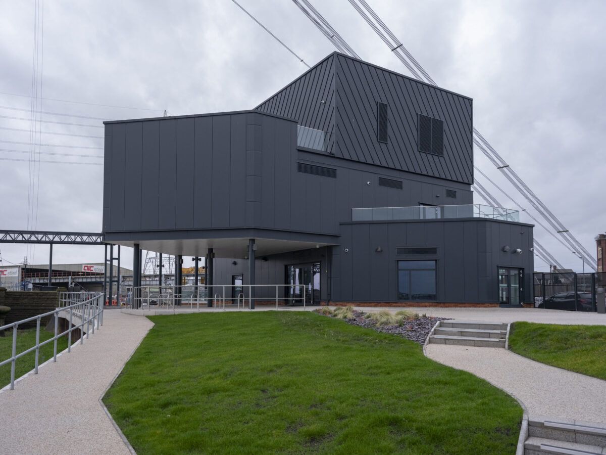The completed visitor centre at Newport Transporter Bridge: dark grey metal cladding, a cantilevered upper floor, and the bridge cables overhead.