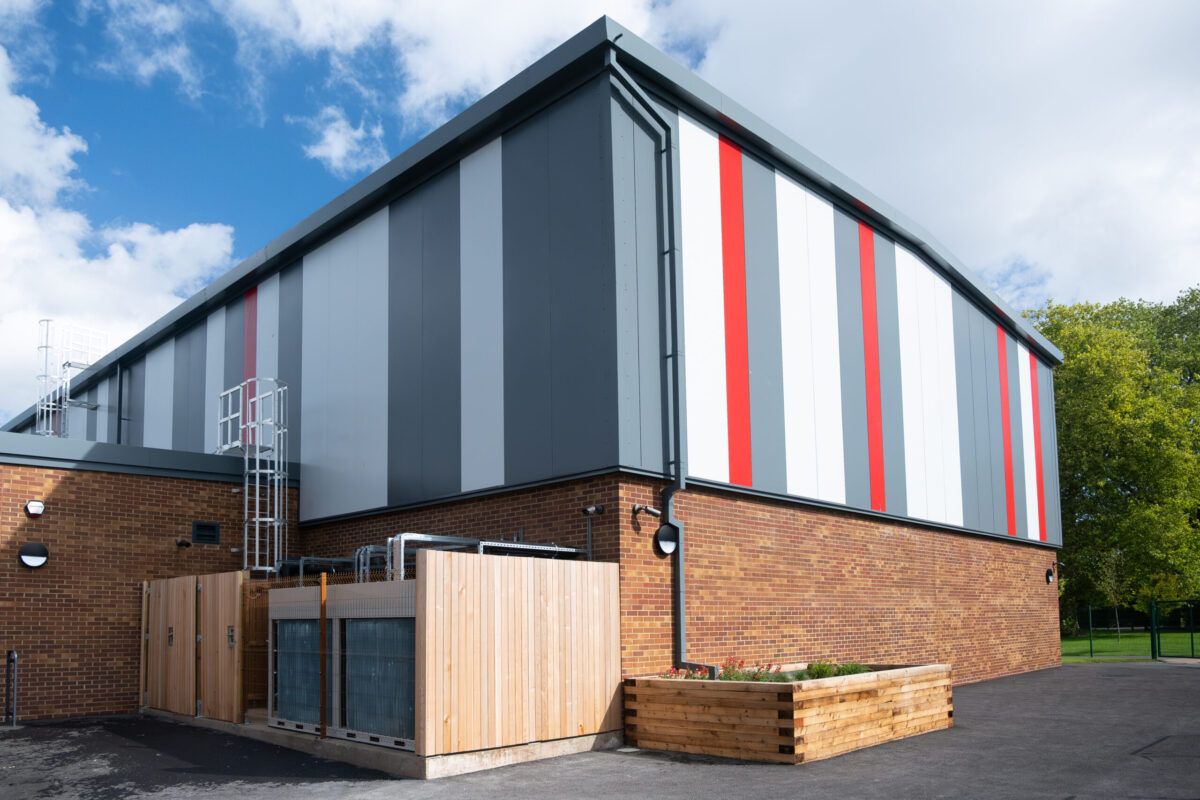 Corner of the sports hall, brick below and tall grey, white and red cladding above, with a timber plant enclosure.
