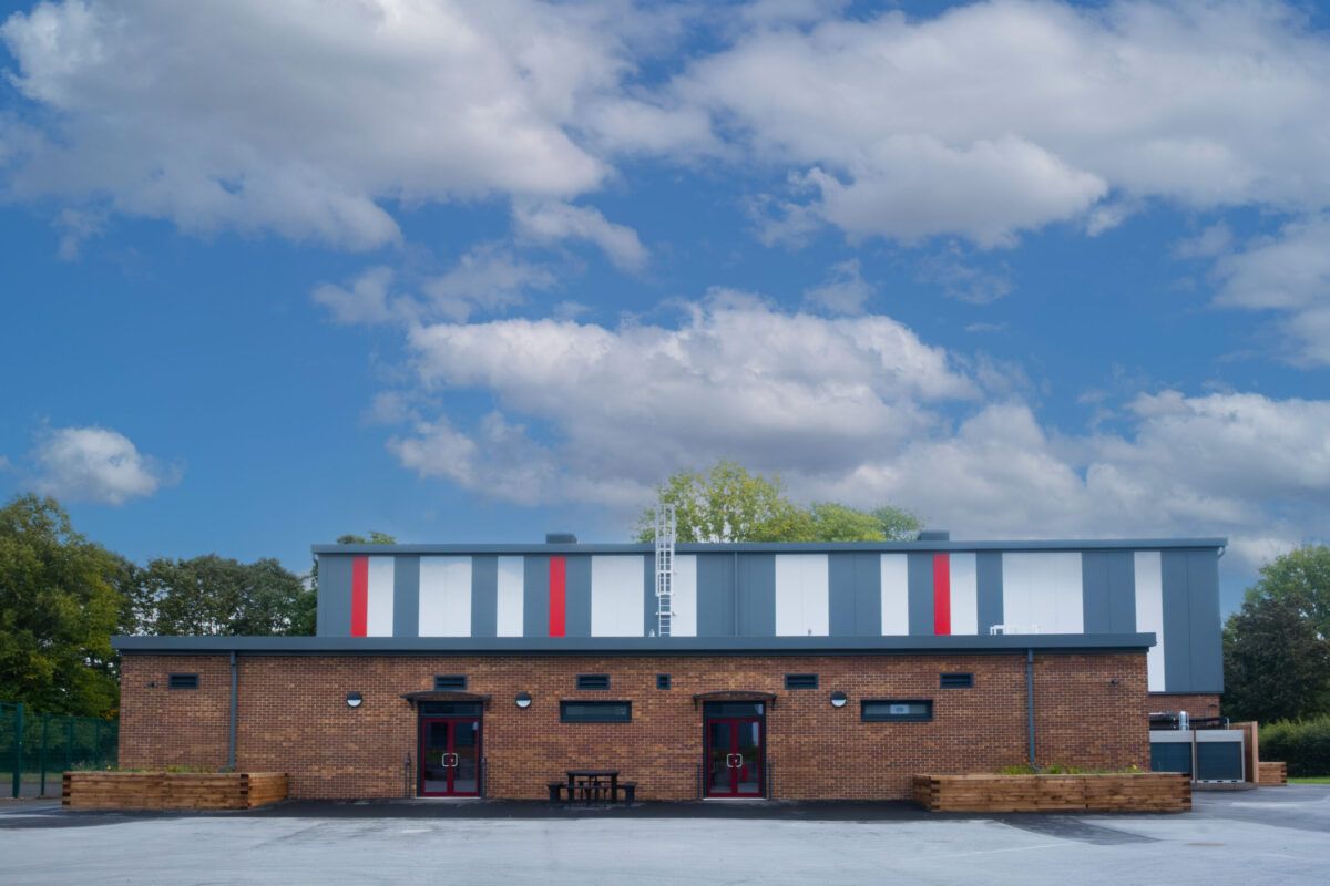 The completed sports hall at Ysgol Gyfun Gwent Is Coed: brick lower storey, grey and white cladding with red stripes, and two red entrance doors.